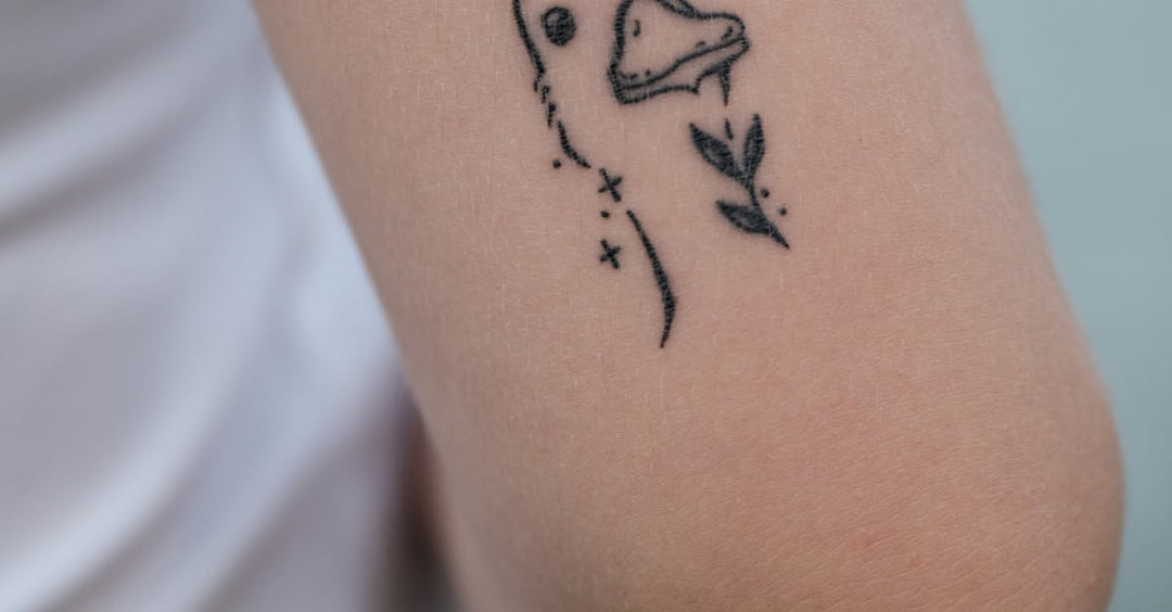 Minimalist micro tattoo of a bird and botanical elements on a person’s upper arm